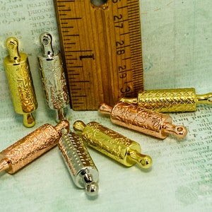 May include: Seven miniature rolling pins in gold, silver, and rose gold finishes. Each rolling pin has a different embossed design. The rolling pins are arranged in a circle on a textured surface.