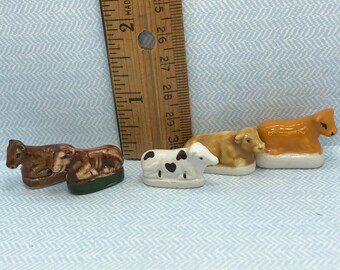 May include: Four miniature ceramic cows in various colors and patterns. The cows are approximately 1 inch tall and are sitting on a white surface with a ruler for scale.