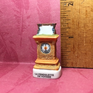 Puede incluir: Un pequeño reloj decorativo de porcelana con un marco de madera marrón y una esfera blanca. El reloj está etiquetado como "LA PENDULETTE DE VOYAGE".
