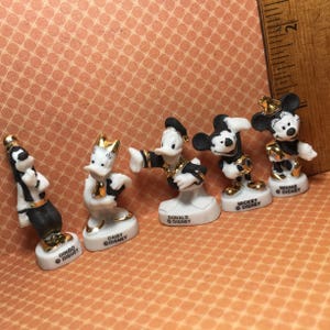 May include: Set of six porcelain Disney character figurines. Each figurine is painted in black and white with gold accents. The characters include Mickey Mouse, Minnie Mouse, Donald Duck, Daisy Duck, Goofy, and Pluto. Each figurine has the character's name and Disney copyright on the base.