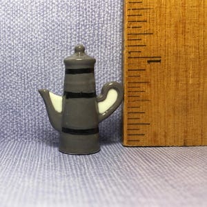 May include: A miniature gray teapot with three horizontal black stripes, a white spout, and a white handle, positioned next to a wooden ruler showing a height of approximately 1.25 inches for the object.