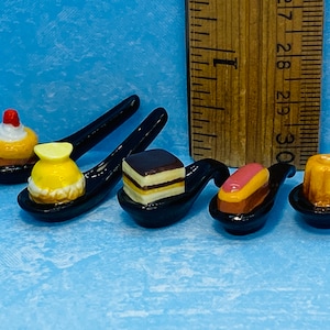 Può includere: Miniature di dessert su cucchiai neri. I dessert includono un cupcake, una pasticceria a forma di limone, una torta a strati, un éclair e un flan. Un righello di legno è sullo sfondo. Questi dessert colorati e dettagliati sono perfetti.