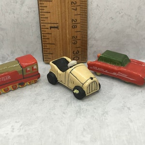 May include: Three miniature ceramic toy vehicles: a red and green train with the word "ECLAIR" on the side, a white car with a black driver, and a red car with a green roof and the words "SOUS MARIN" and "Dinette" on the side.