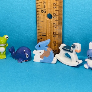 Puede incluir: Colección de figuras de cerámica en miniatura, que incluyen una rana verde, una ballena azul, un conejo azul y marrón, un caballito balancín blanco y un conejo azul. Una regla de madera está en el fondo. Las figuras están sobre una superficie azul.