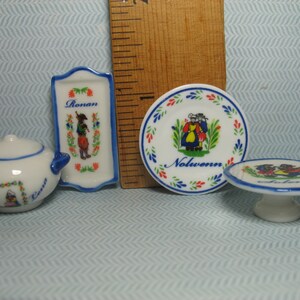 May include: Set of four miniature ceramic dishes with blue and white trim. The dishes feature a traditional folk art design with people in colorful clothing. The dishes are labeled "Rouen" and "Nolwenn".