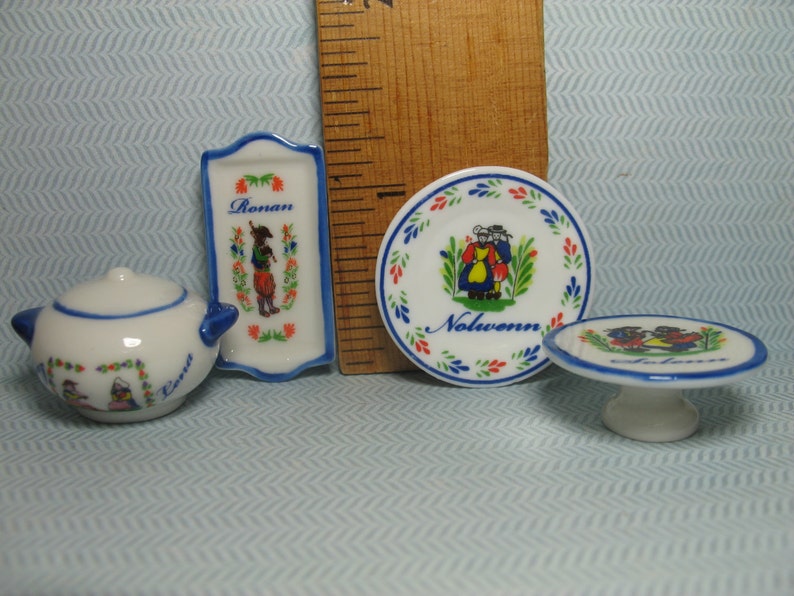 May include: Set of four miniature ceramic dishes with blue and white trim. The dishes feature a traditional folk art design with people in colorful clothing. The dishes are labeled "Rouen" and "Nolwenn".
