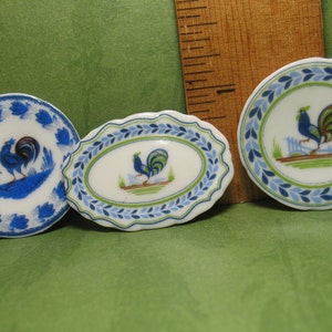 May include: Three miniature ceramic plates with blue and green floral designs and a rooster illustration. The plates are white with blue and green accents.