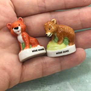 Pode incluir: Duas pequenas estatuetas de cerâmica de uma raposa e um urso. A raposa é laranja e branca e o urso é marrom. Ambas as estatuetas estão sentadas em uma base verde e têm as palavras "Bébé Renard" e "Bébé Ours" escritas na base.