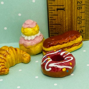 Puede incluir: Un conjunto de cuatro pasteles de cerámica en miniatura, que incluyen un croissant, una dona con glaseado rosa y chispas, una crema de hojaldre con glaseado rosa y blanco y un eclair largo con glaseado de chocolate.