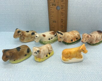 May include: A collection of seven miniature ceramic figurines of cows and a dog. The figurines are painted in brown, black, white, and yellow with black spots. The figurines are all lying down.