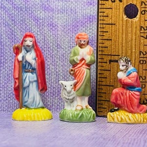 May include: Three small porcelain figurines depicting the nativity scene. A shepherd with a staff and red robe, a man with a green robe and a white lamb, and a kneeling man with a red robe.