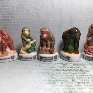 Peut inclure: Cinq figurines miniatures en céramique représentant différentes espèces de singes, chacune avec une couleur et une pose distinctes. Les figurines sont présentées sur un fond blanc avec le nom de chaque espèce écrit en dessous : Orang-Outan, Talapoin, Macaque Japonais, Gorille et Langur Douc Male.