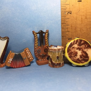 May include: A set of five miniature musical instruments, including a harp, an accordion, a lyre, a drum, and a tambourine. The instruments are made of ceramic and are painted in various shades of brown, yellow, and white.