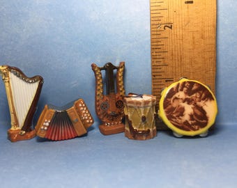 May include: A set of five miniature musical instruments, including a harp, an accordion, a lyre, a drum, and a tambourine. The instruments are made of ceramic and are painted in various shades of brown, yellow, and white.