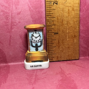 May include: A miniature clock figurine with a gold-colored base and top, and a white face with black numbers. The clock face is surrounded by a light blue frame. The base has the text "LE CARTEL".