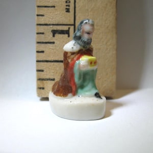 Puede incluir: Una figura de porcelana en miniatura de un personaje arrodillado con barba, vestido con una túnica marrón y verde, que sostiene un pequeño objeto amarillo. La figura está junto a una regla de madera que muestra una altura de 3,2 centímetros.