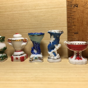 May include: Five miniature porcelain vases in various styles and colors. The vases are decorated with gold trim and floral designs. The vases are arranged in a row on a wooden surface.