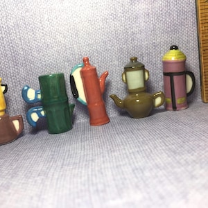 May include: A collection of six miniature ceramic coffee pots in various colors and designs. The pots are all different shapes and sizes, and some have handles.