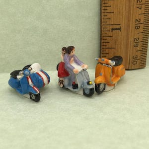 May include: Three miniature ceramic scooter figurines next to a wooden ruler showing a scale of one inch. One blue scooter with white stripes, one light blue scooter with two figures, and one orange scooter, each with black seats and wheels.