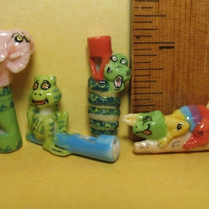 Op de afbeelding: Vier keramische fluitjes met dierendesigns. Een roze olifant, een groene kikker, een groene slang en een groen-gele hagedis.