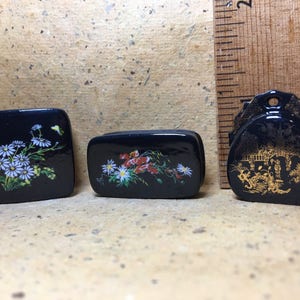 Può includere: Tre piccole scatole nere con accenti dorati e disegni floreali. Le scatole sono decorate con fiori bianchi e rossi, e una scatola ha un disegno dorato di un edificio.