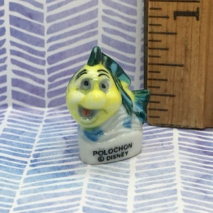 May include: A small ceramic figurine of a yellow and blue fish with a smiling face. The figurine is labeled "Polochon Disney".