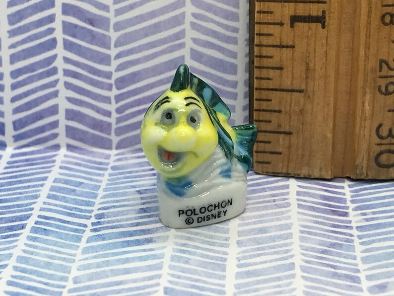 May include: A small ceramic figurine of a yellow and blue fish with a smiling face. The figurine is labeled "Polochon Disney".