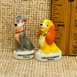 Puede incluir: Dos figuras de perro de cerámica, una gris y otra amarilla con orejas rojas, cada una sobre una base blanca con la inscripción LA BELLE ET LE CLOCHARD 1955 Disney. Una regla de madera al lado muestra una altura de unos 6,5 cm.