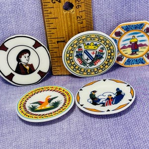 May include: Five miniature ceramic plates with various designs. One plate features a portrait of a person in a red and white outfit, another has a coat of arms with a red cross, one has a bird with yellow and orange feathers, one has a family of three people, and the last one has a person in a blue and white outfit.