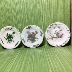 Puede incluir: Tres platos pequeños de cerámica blanca con diseños florales verdes, rosas y amarillos. Los platos están sobre un fondo verde.