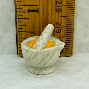 May include: A miniature white mortar and pestle with a grey marble-like pattern, containing a textured orange substance. The item is positioned in front of a wooden ruler, showing a height of approximately 1 inch for the mortar bowl.