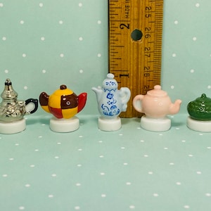 May include: Set of six miniature ceramic teapots in various colors and patterns, including silver, red, yellow, blue, pink, and green. The teapots are displayed on a white surface.