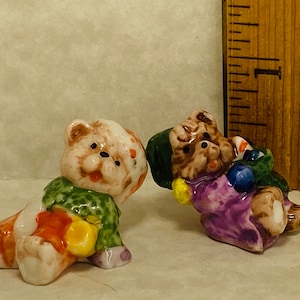May include: Two small ceramic bear figurines. One bear is wearing a green and orange outfit and the other is wearing a purple and green outfit. Both bears have brown fur and are holding colorful objects.