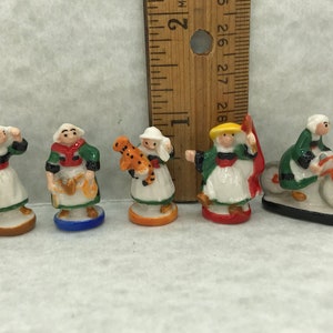 May include: Five miniature figurines of people wearing traditional clothing. The figurines are painted in bright colors and have detailed features. The figurines are standing on small bases. The figurines are all different and represent different cultures.