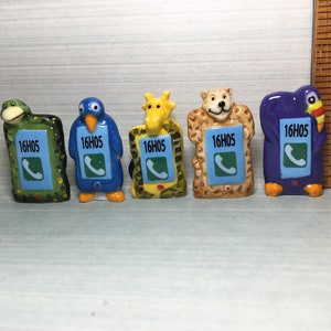 May include: Five ceramic figurines of different animals, each holding a small blue phone with the text "16H05" on the screen. The animals are a frog, a penguin, a giraffe, a bear, and a toucan.