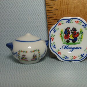 May include: A white porcelain teapot and saucer set with a blue and red floral design. The saucer has a scene of two people in traditional clothing and the text "Morgan".
