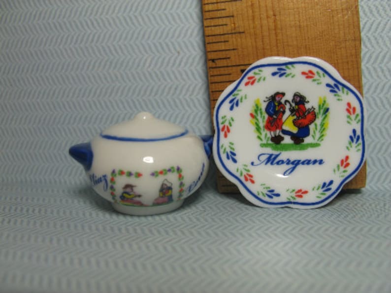 May include: A white porcelain teapot and saucer set with a blue and red floral design. The saucer has a scene of two people in traditional clothing and the text "Morgan".