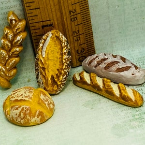Puede incluir: Surtido de panes en miniatura, que incluye una hogaza, una baguette y una espiga de trigo. Los panes están pintados con detalles marrones y blancos. Una regla de madera está en el fondo. Los panes miden unos 2-3 cm de largo.