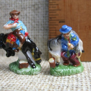 May include: Two porcelain figurines depicting cowboys on horseback. The cowboy on the left is wearing a blue shirt, brown hat, and red bandana. The cowboy on the right is wearing a blue jacket, blue hat, and red trousers. Both figurines are standing on a green base.