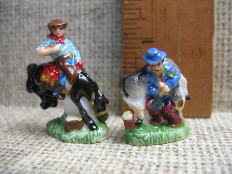 May include: Two porcelain figurines depicting cowboys on horseback. The cowboy on the left is wearing a blue shirt, brown hat, and red bandana. The cowboy on the right is wearing a blue jacket, blue hat, and red trousers. Both figurines are standing on a green base.