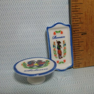 May include: Two small ceramic plates with blue and white trim. One plate has a floral design and the word "Ronan" printed on it. The other plate has a blue and white design with two figures and the word "Solan" printed on it.