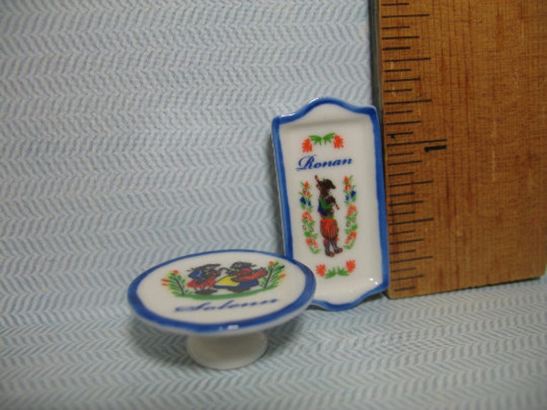 May include: Two small ceramic plates with blue and white trim. One plate has a floral design and the word "Ronan" printed on it. The other plate has a blue and white design with two figures and the word "Solan" printed on it.