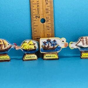 May include: Four miniature ceramic bottle-shaped figurines with a nautical theme. Each bottle features a different ship with sails and a blue and white ocean scene. The bottles are decorated with gold trim and the text "Les Bateaux de la Mer".