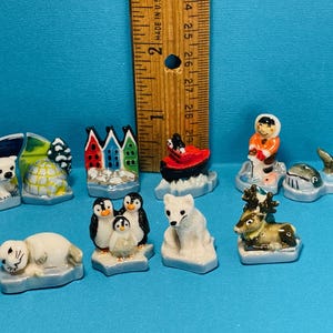 May include: A set of 10 miniature ceramic figurines depicting various arctic animals and scenes. The figurines include a polar bear, a seal, a reindeer, a penguin, a walrus, a boat, a house, and an igloo. The figurines are painted in bright colors and have a whimsical style.