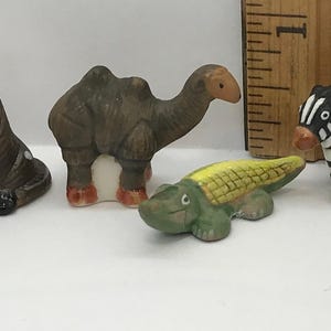 May include: A miniature brown camel figurine and a small green and yellow crocodile figurine positioned next to a wooden ruler showing a measurement of approximately 1 inch. The camel has two humps and the crocodile has a textured back.