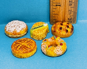 May include: Five miniature ceramic pastries, including a round pastry with white icing, a round pastry with a lattice crust, a ring-shaped pastry with white icing, a round pastry with a four-leaf clover design, and a pastry with a yellow and green design that says "Colombier".