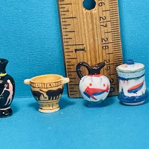 May include: Four miniature ceramic vases with painted designs. One vase is black with yellow and pink details, another is white with red and blue details, another is brown with black details, and the last is white with blue and green details.