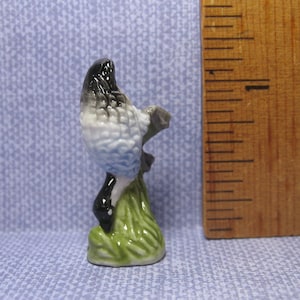 Puede incluir: Una pequeña figurita de cerámica de un pájaro azul y blanco posado en una rama verde con hojas. El pájaro tiene la cabeza negra y el cuerpo blanco con detalles azules.
