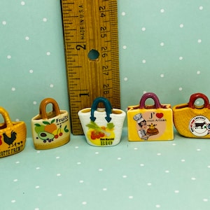 May include: Five miniature ceramic tote bags with different designs. The bags are yellow, white, and brown with various images and text. The bags are approximately 1 inch tall.