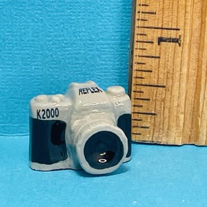 May include: A gray and black miniature camera with the words "K2000" and "REFLEX" printed on it.